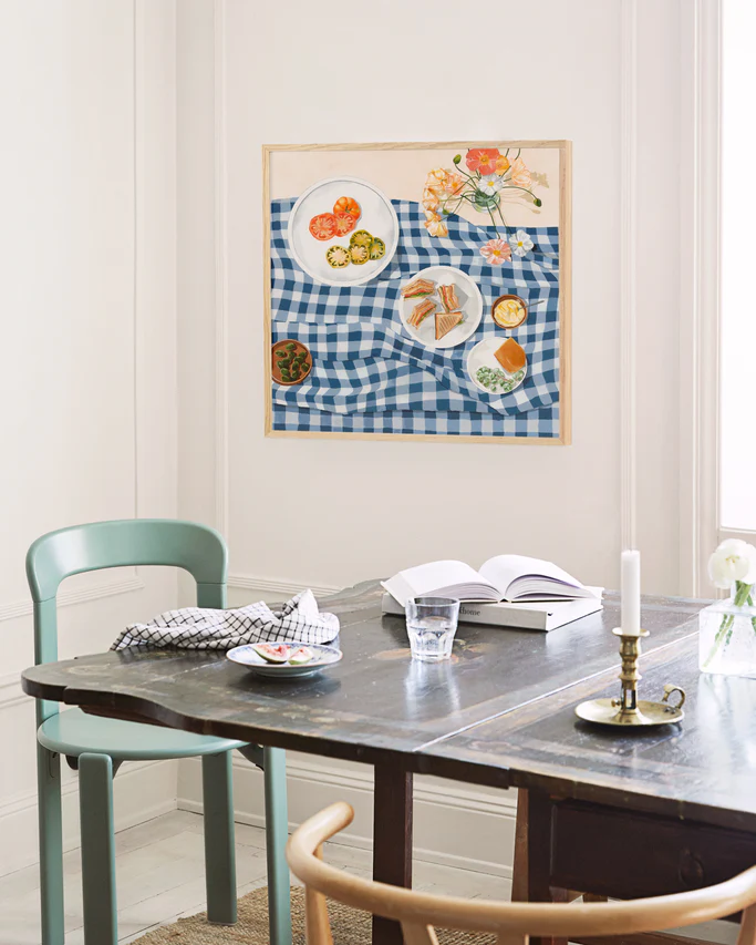 A kitchen with a dark wood table. On the table are two books, a water glass, a vase with white flowers, a golden candlestick, a small plate with figs, and a checkered kitchen towel. Above the table hangs an oak-framed poster titled "Poppies and Picnics."