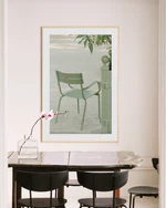 The Asseyez-vous Poster, depicting a green chair, is displayed on a white wall above a wooden table accompanied by dark chairs. A glass vase with an elegant orchid graces the table, enhancing the minimalist rooms elegance.