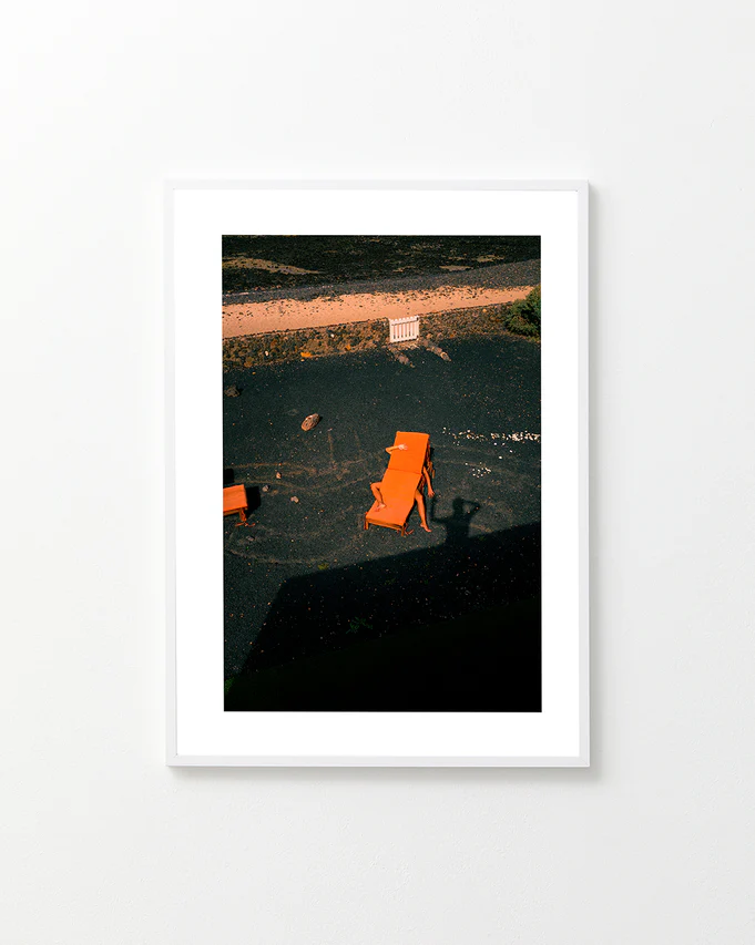 The Short Romantic Story Between a Chair and a Shadow Poster features three orange lounge chairs creating captivating shadows on black sand under bright light, with two more chairs by a white fence in the background, all viewed from an elevated angle on a tranquil beach.