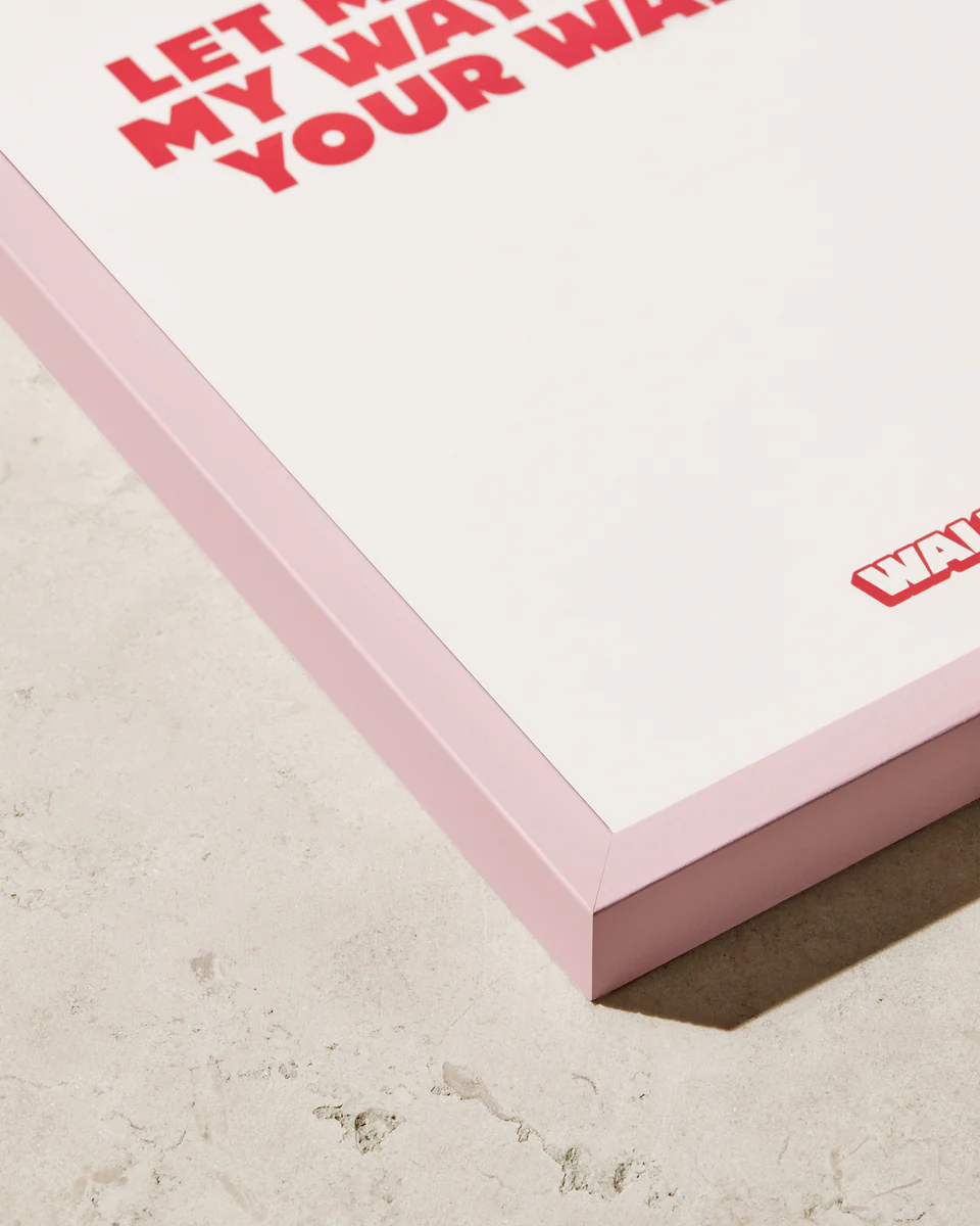 The image showcases the Frame Pastel Pink, a framed artwork with a pastel pink wooden frame that adds stylish color. It features prominent red text and is placed on a light-textured surface.