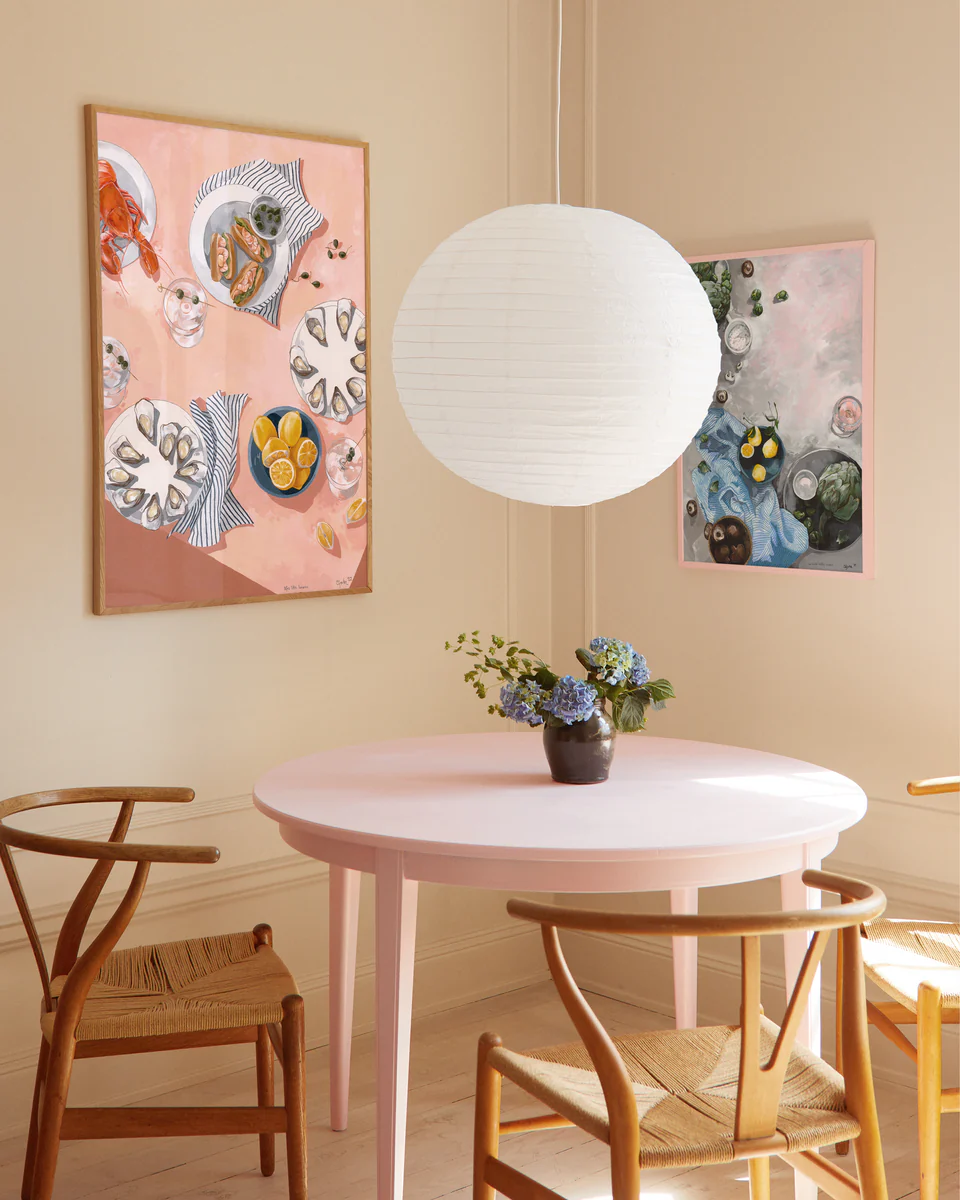 A cozy room boasts a pink round table with wooden chairs, a vase of blue flowers at its center. The walls feature the Lifes Little Luxuries poster with dining and plates themes. Overhead, a large white paper lantern completes the scene.