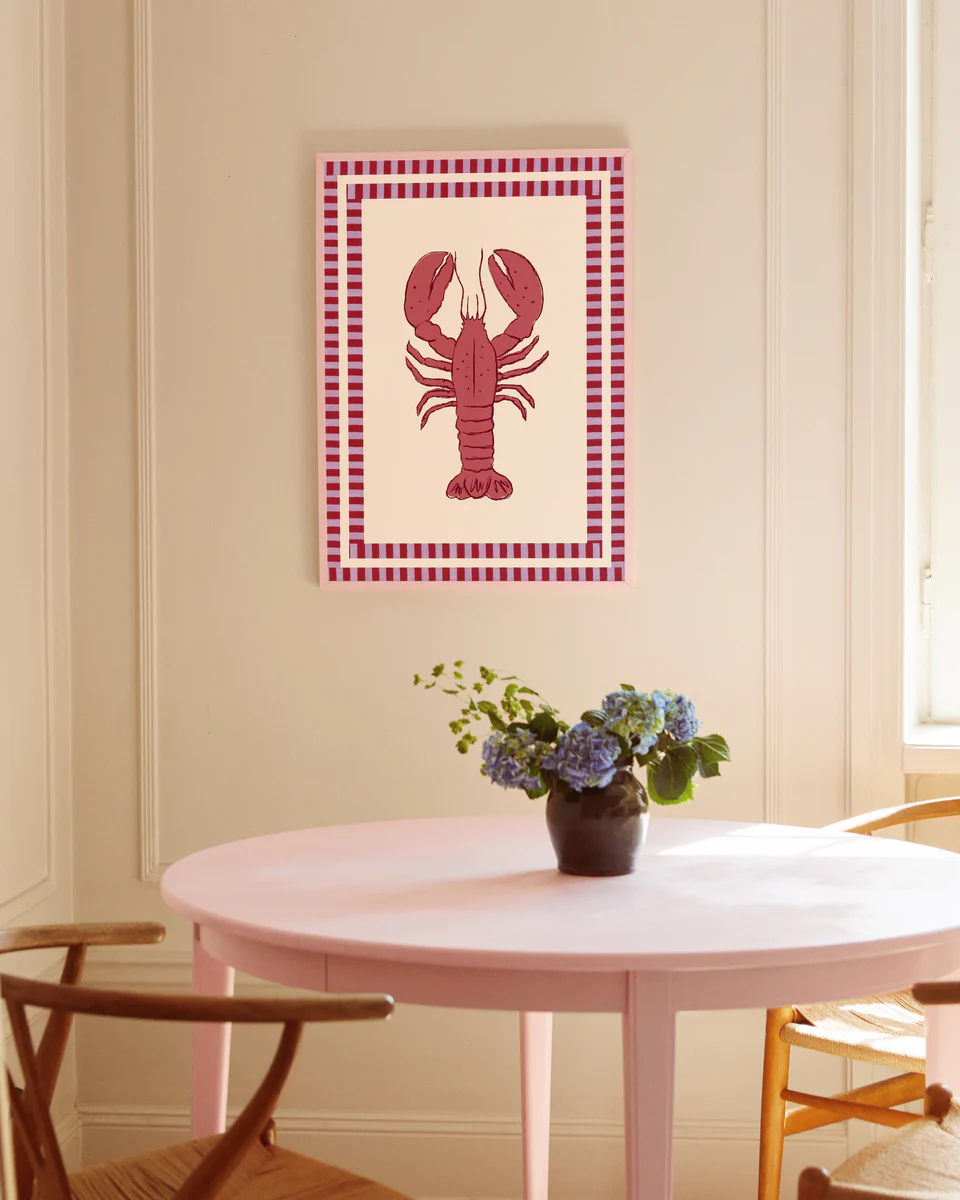 A dining area features a round pink table with wooden chairs and a small vase of purple and blue flowers. The beige wall displays the framed Le Stripy Homard Poster, an artwork with a checkered border showcasing a red lobster collage. Sunlight streams in from a nearby window.