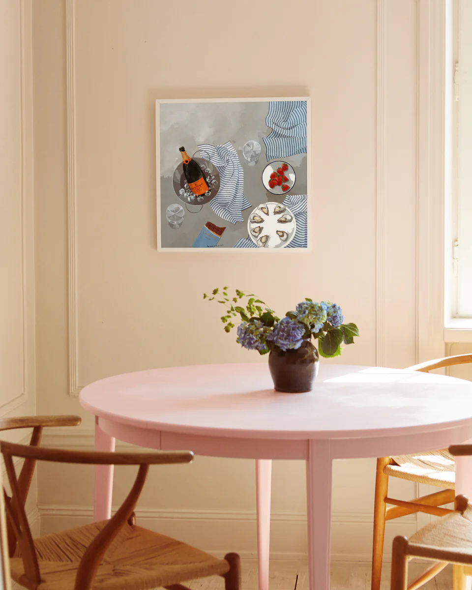 A cozy room with a round pink table and wooden chairs features a small vase of blue hydrangeas and a bottle of Veuve Clicquot. On the cream-colored wall, The Veuve Made Me Do It poster complements the soft light streaming through the window.