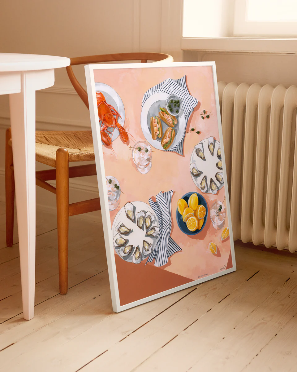 The framed Lifes Little Luxuries Poster captures an indulgent dining scene with oysters, lemons, bread, and a lobster. Its peach background complements the setting beside a wooden chair and white radiator on light wood floors, celebrating seafood delicacies in style.