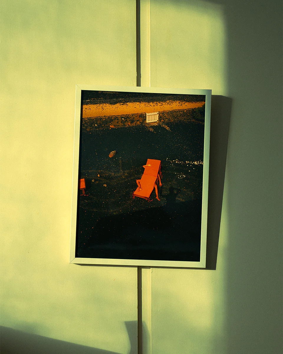 The Short Romantic Story Between a Chair and a Shadow Poster depicts a red-orange chair casting long shadows on a dark surface, reminiscent of a tranquil beach. A sandy area and small white fence enhance the backdrop, while sunlight creates gentle patterns on the greenish wall.