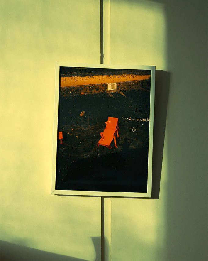 The Short Romantic Story Between a Chair and a Shadow Poster depicts a red-orange chair casting long shadows on a dark surface, reminiscent of a tranquil beach. A sandy area and small white fence enhance the backdrop, while sunlight creates gentle patterns on the greenish wall.