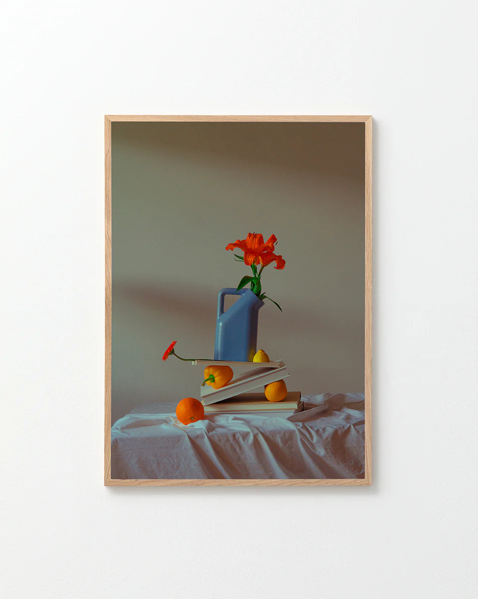 The Hanging in There Poster features a vibrant scene: a blue watering can with an orange flower on open books, surrounded by two oranges and a lemon on white cloth, evoking Berlins artistic charm against a simple backdrop.