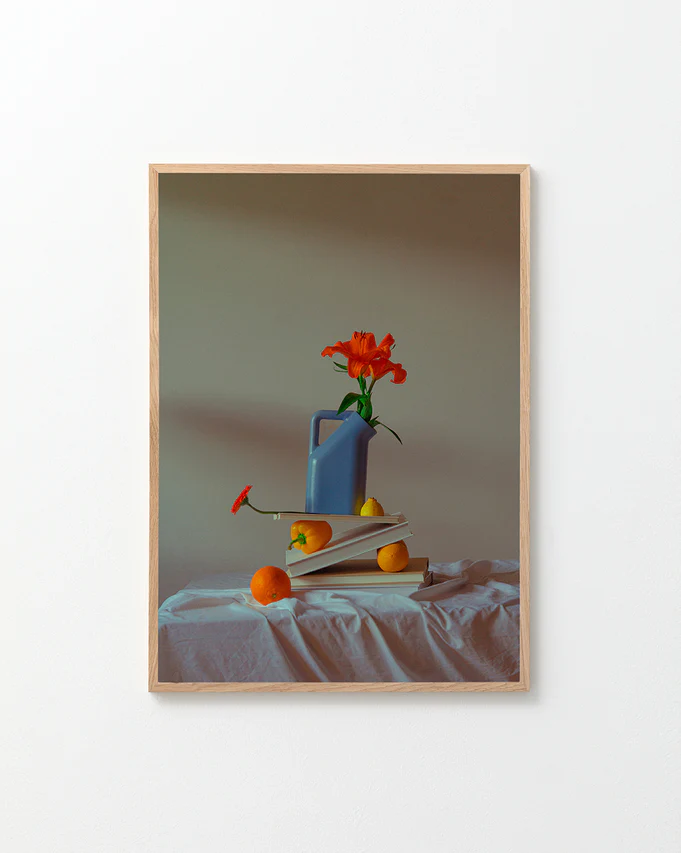 The Hanging in There Poster features a vibrant scene: a blue watering can with an orange flower on open books, surrounded by two oranges and a lemon on white cloth, evoking Berlins artistic charm against a simple backdrop.