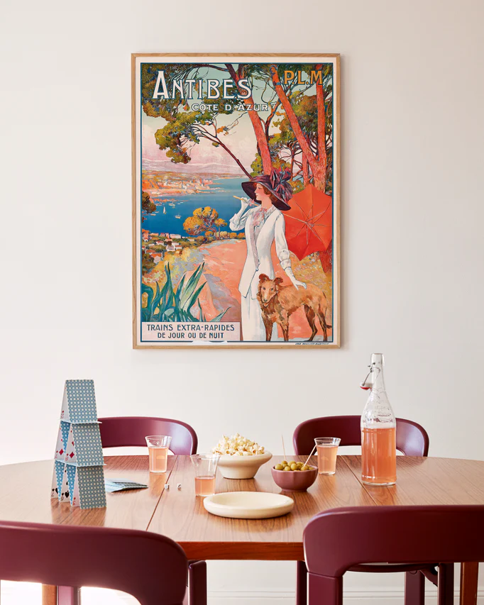 A cozy dining area showcases a round wooden table with three red chairs, topped with popcorn, drinks, and a deck of cards. The white wall behind is adorned with the Antibes Cote dAzur Poster, bringing the French Rivieras colorful charm to the space.