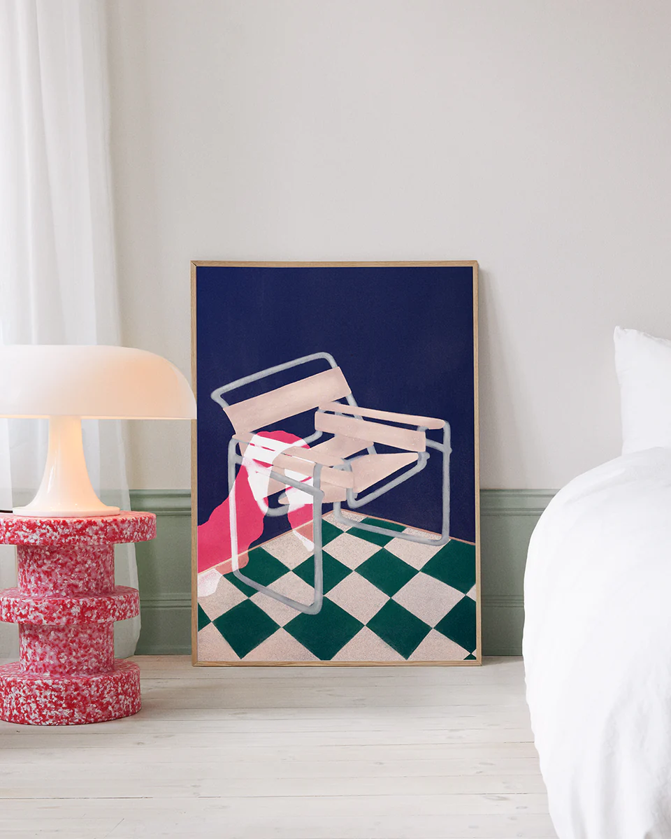 A minimalist room showcases the Bauhaus 02 Poster with a framed abstract of a white Wassily chair over a red and green checkered floor, set against a dark blue backdrop. The artwork leans beside a modern lamp on a bold red table, capturing Bauhaus modernisms essence.