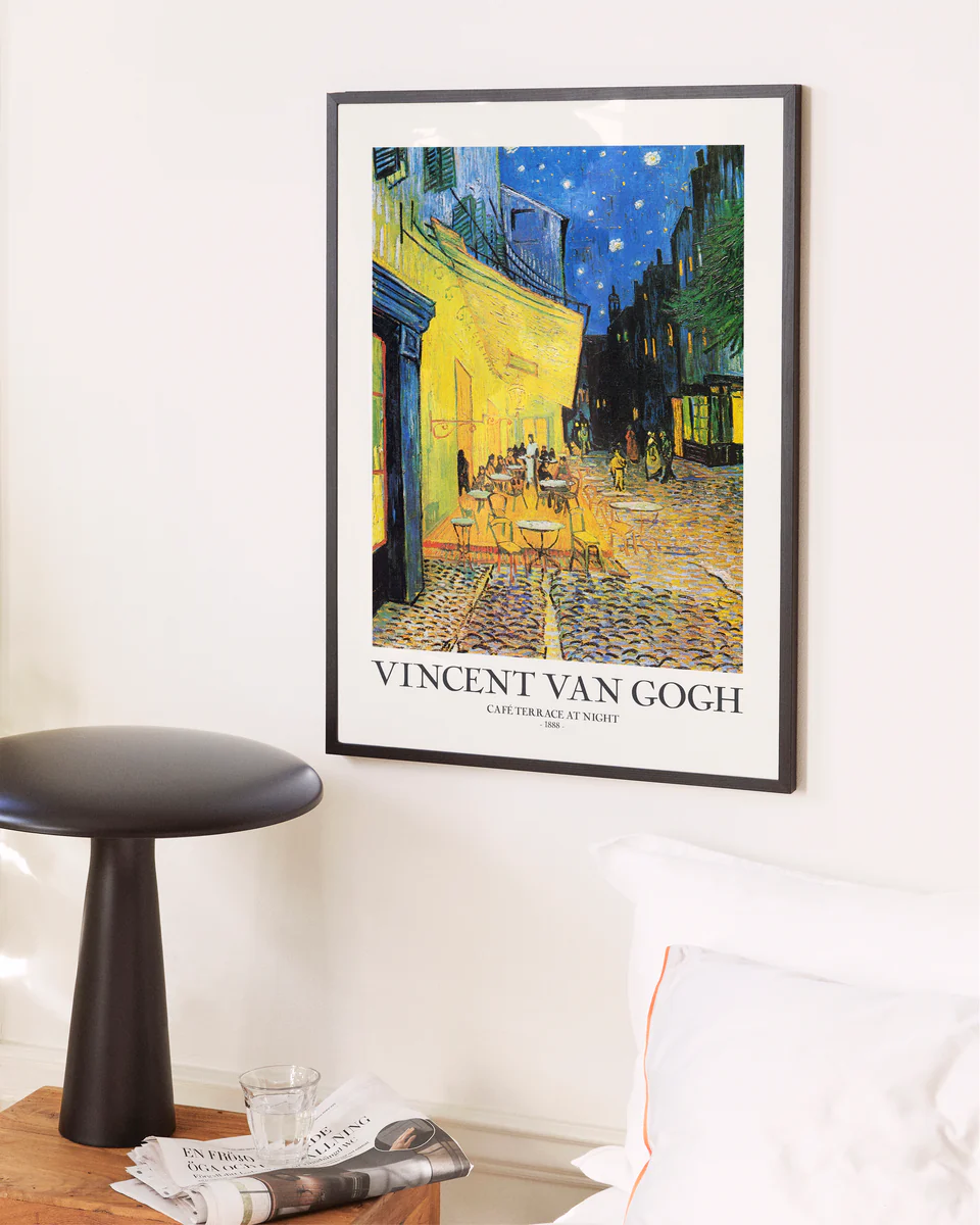 Café Terrace at Night by Vincent van Gogh Poster framed on a white wall, depicting a vibrant café scene under a starry evening sky. Below it, the artists name and title are displayed. Nearby, a small bedside table holds magazines and a glass.