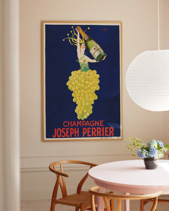 The room features a Champagne Joseph Perrier Poster framed on the wall, depicting a grape-made figure holding a champagne bottle against a blue backdrop, complemented by a wooden chair, round table with blue flowers in a vase, and a hanging white paper lantern.