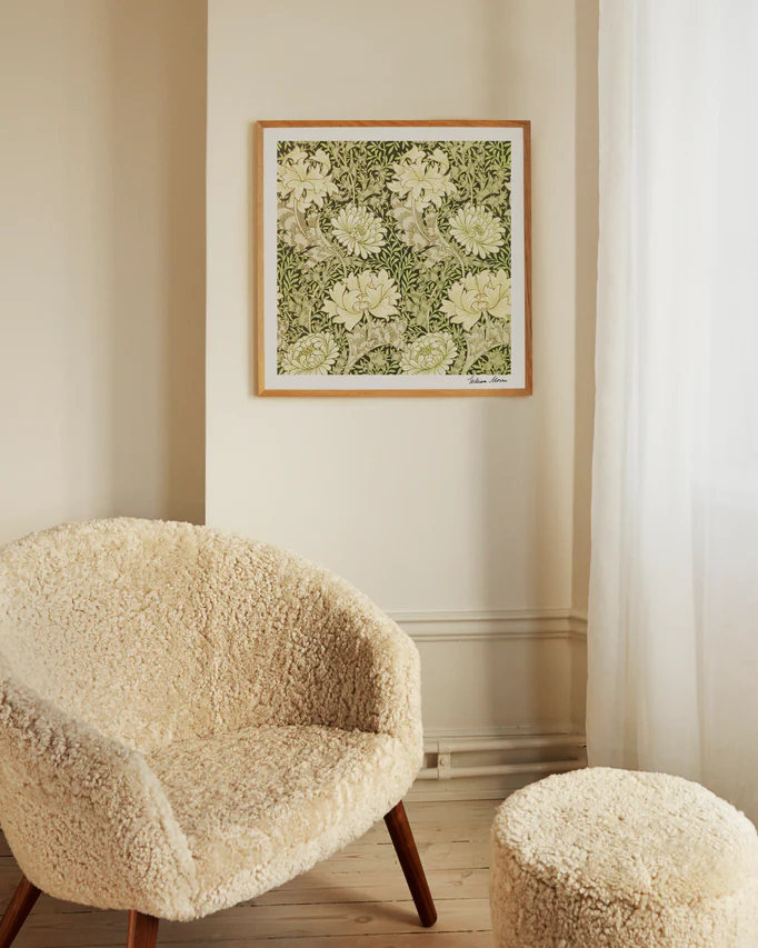 A cozy room features a textured armchair and footstool with wooden legs. The wall showcases the Chrysanthemum Pattern by William Morris poster. Soft light through white curtains enhances the warm and inviting ambiance.