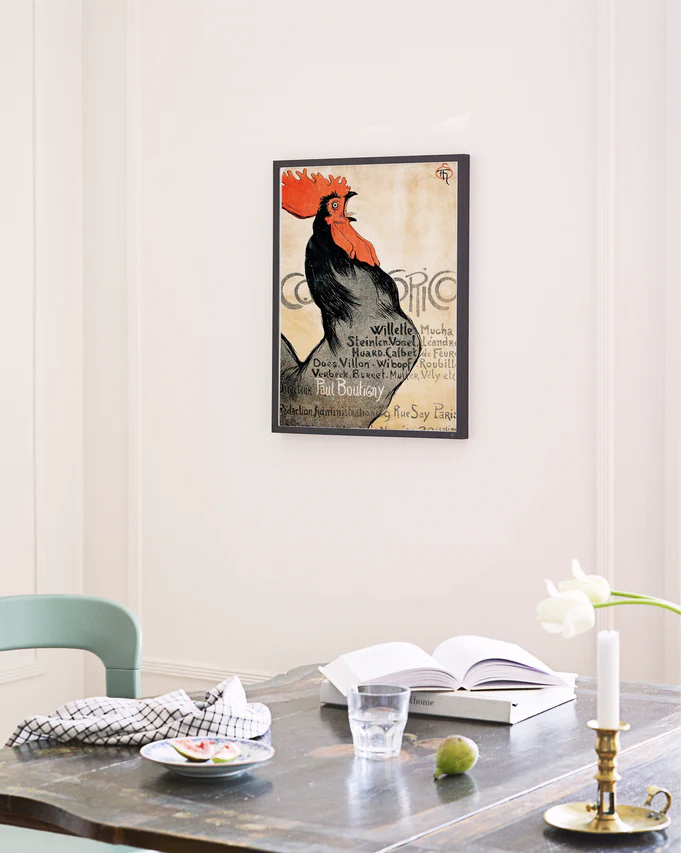 A Cocorico by Théophile-Alexandre Steinlen poster adds vintage charm to the wall. Below, a wooden table holds an open book, a glass of water, and a black-patterned white napkin. In front, a white tulip in a glass vase embodies French Art Deco elegance.