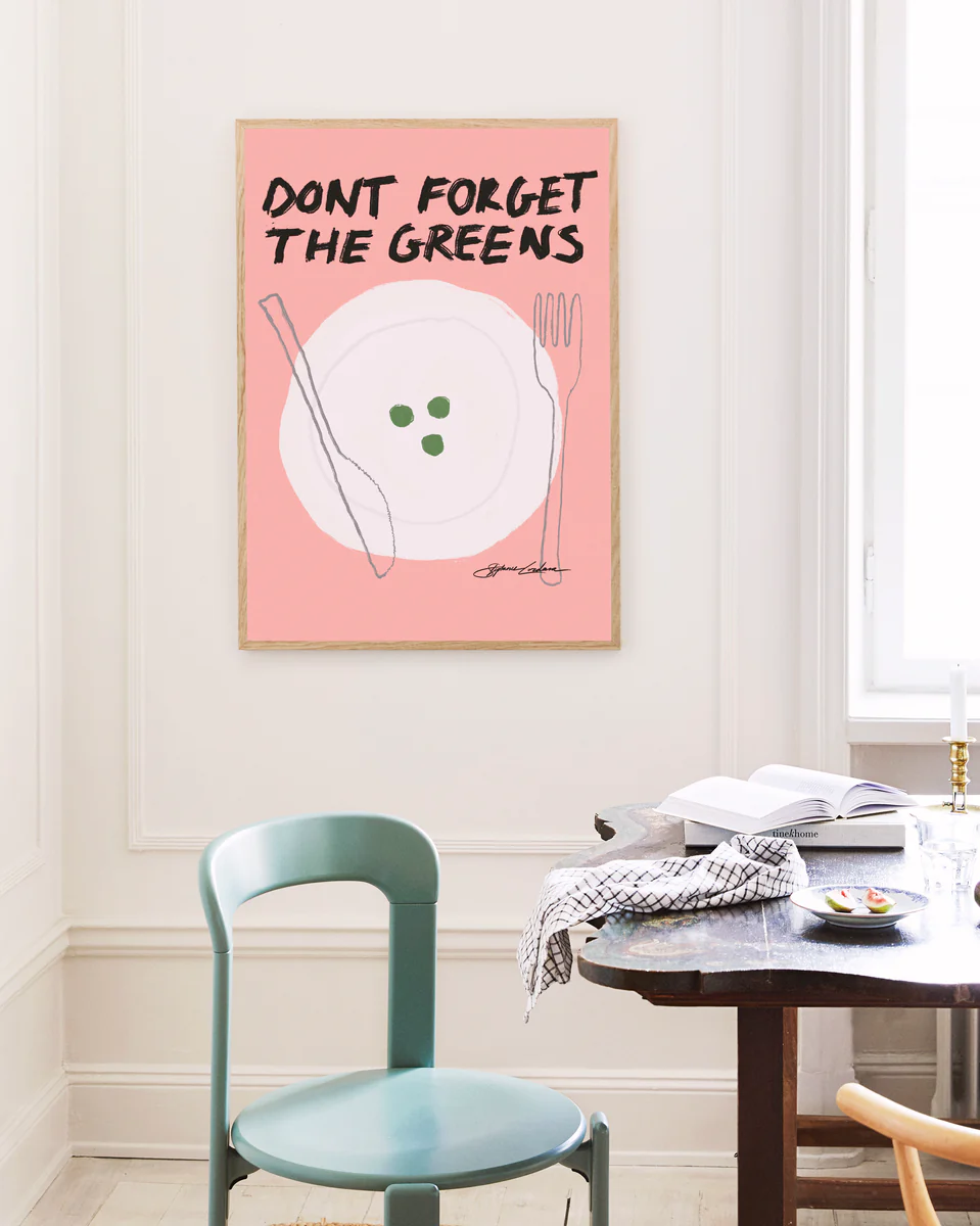 In a minimalistic dining room, a turquoise chair stands next to a dark wooden table. A humorous poster titled Dont Forget the Greens on the wall, with its pink backdrop and quirky cutlery and peas illustration, adds charm and whimsy to the tasteful decor.