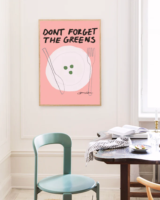 In a minimalistic dining room, a turquoise chair stands next to a dark wooden table. A humorous poster titled Dont Forget the Greens on the wall, with its pink backdrop and quirky cutlery and peas illustration, adds charm and whimsy to the tasteful decor.
