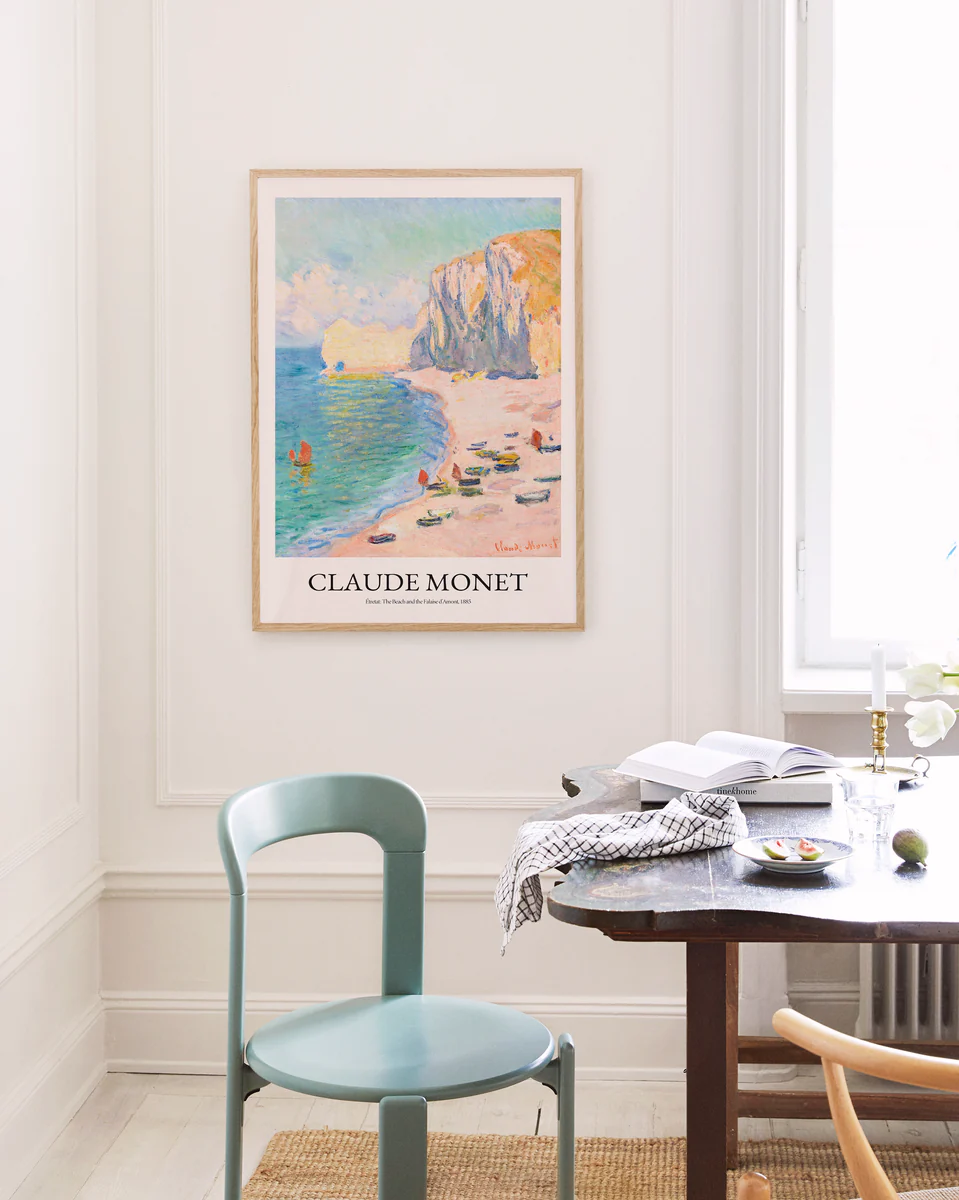A serene room with a light green chair and wooden table exudes French elegance. The Étretat: The Beach and the Falaise dAmont by Claude Monet poster adorns the wall. An open book, glasses, plates, and a candle sit on the table as natural light enhances this tranquil French Riviera-inspired setting.