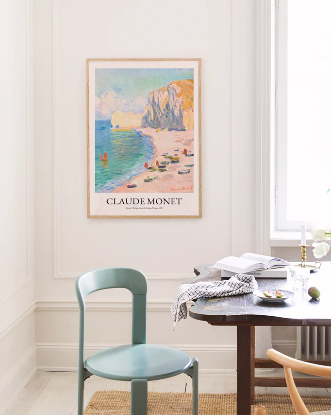 A serene room with a light green chair and wooden table exudes French elegance. The Étretat: The Beach and the Falaise dAmont by Claude Monet poster adorns the wall. An open book, glasses, plates, and a candle sit on the table as natural light enhances this tranquil French Riviera-inspired setting.