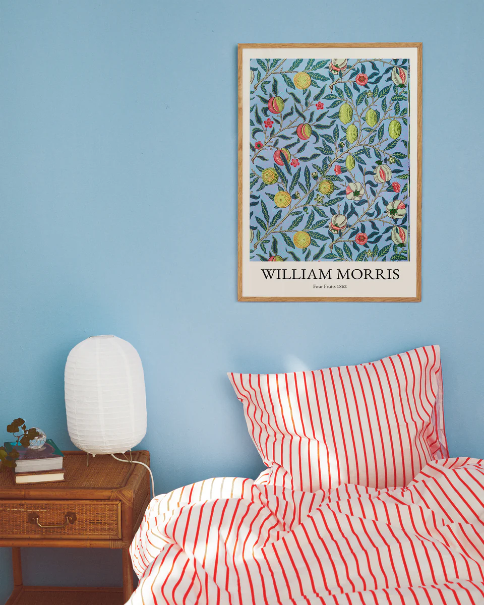 A cozy bedroom with a blue wall displays a Four Fruits by William Morris Poster. The bed has red and white striped bedding, and a wooden side table supports a white textured lamp and books, adding to the rooms charm.