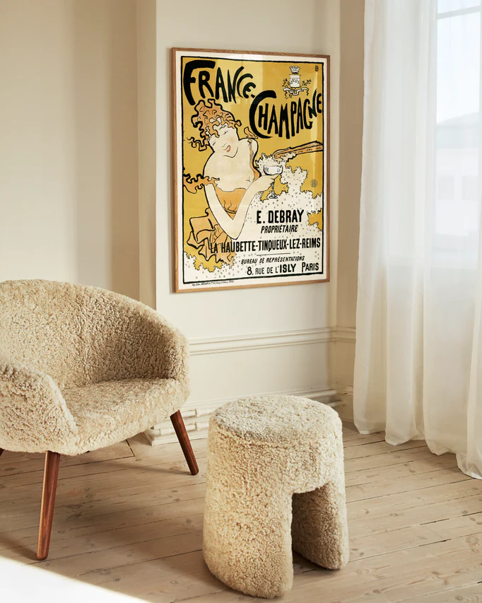 A cozy room showcases a fluffy beige chair and matching stool on a light wooden floor. The wall features the stylish France-Champagne Poster, adding a bold touch. Sheer curtains gently filter soft light from the window, creating an inviting ambiance.