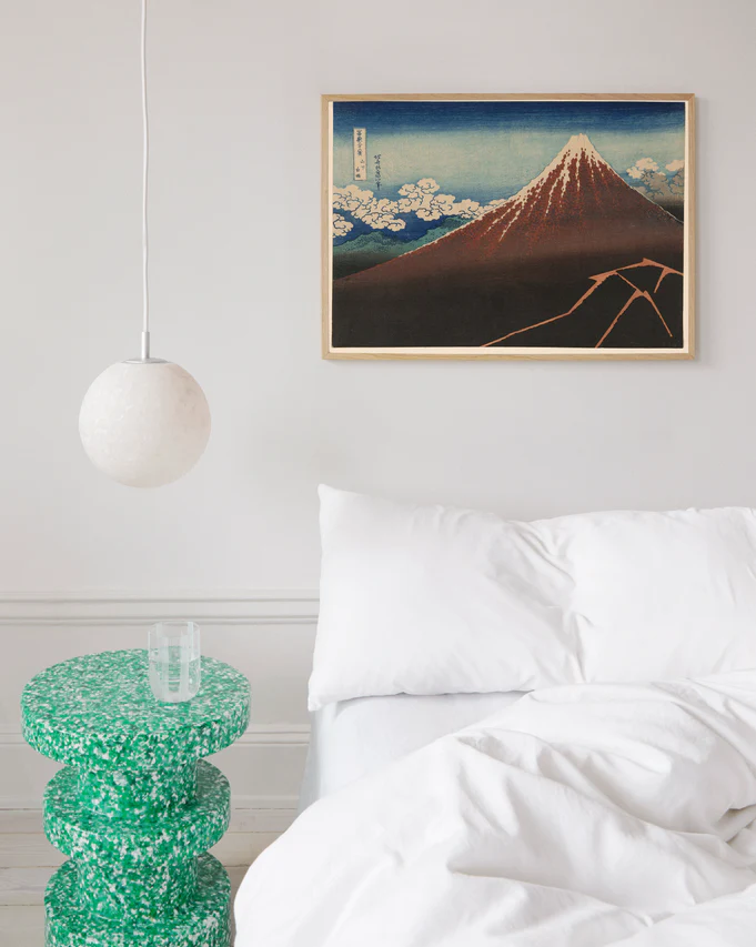 A minimalist bedroom has a white bed with white bedding, a green speckled side table with a glass, and a round white pendant light. A blue and red toned framed poster titled Fuji above the Lightning by Katsushika Hokusai hangs above.