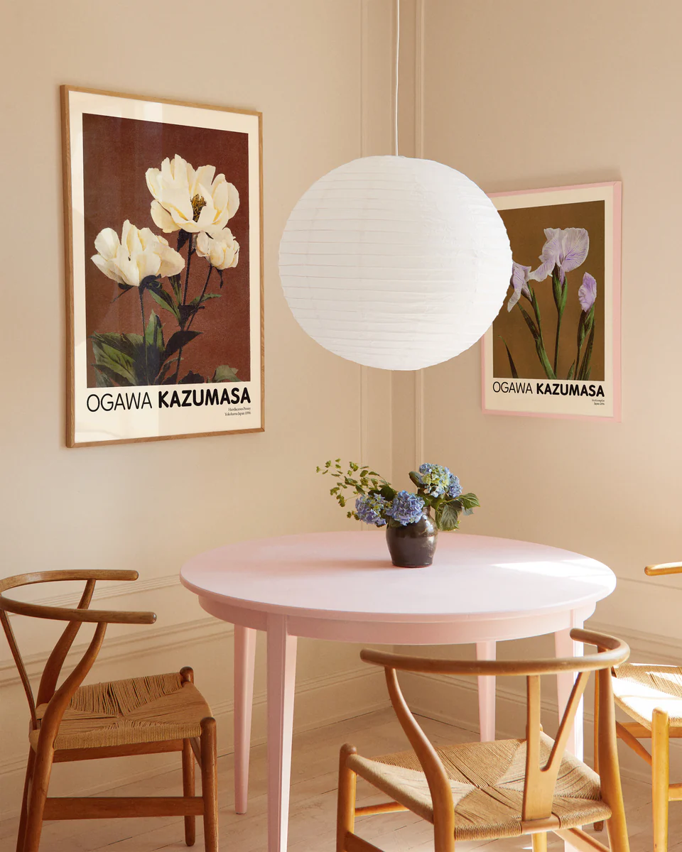 A cozy dining area with a round pink table and four wooden chairs exudes Japanese elegance, featuring a small vase of pale yellow peonies. Two Herbaceous Peony by Ogawa Kazumasa posters enhance the ambiance, illuminated by a white paper lantern above.