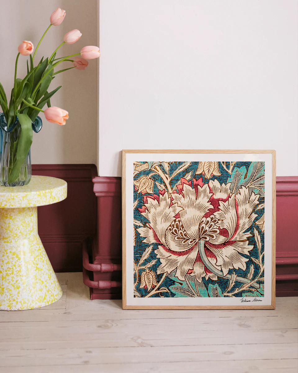 The Honeysuckle by William Morris poster, with intricate floral patterns from the Arts and Crafts era, leans against a wall. Nearby, pink tulips fill a vase on a speckled yellow and white side table. The wall and molding contrast in white and burgundy hues.