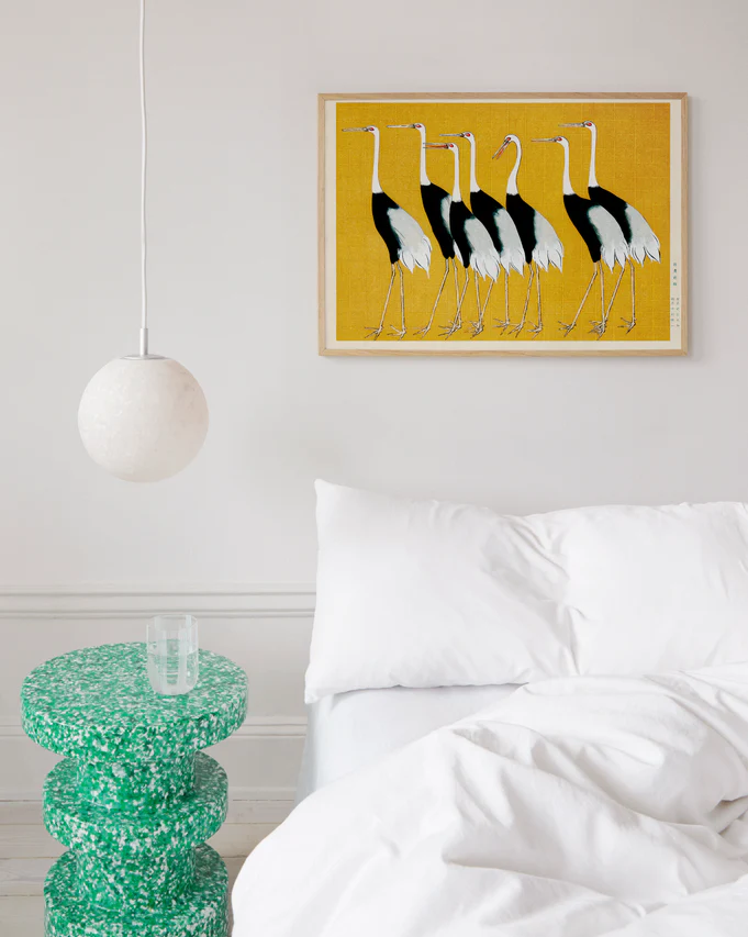 A minimalist bedroom features a white bed, a textured green side table, and a round pendant light. A framed Japanese Red Crown Cranes by Ogata Korin poster on the wall adds vibrant contrast to the neutral decor.