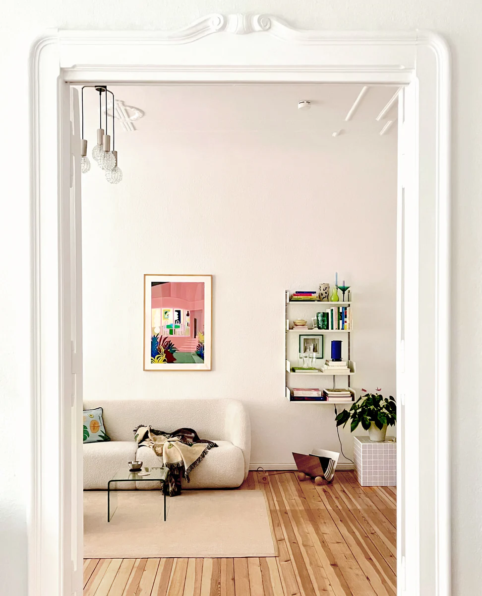 A cozy living room showcases a white sofa with colorful cushions and a throw blanket. The Lighted Windows 01 Poster hangs above a modern coffee table. A potted plant, wall-mounted shelf with books, and decor complement the ambiance, all set against light wood flooring.