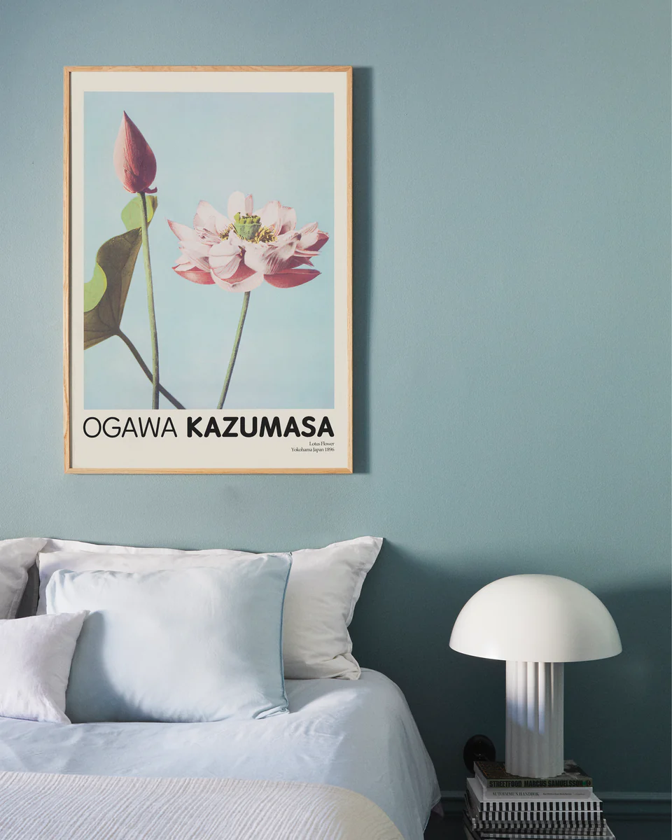 A minimalist bedroom with a light blue wall showcases the Lotus Flowers by Ogawa Kazumasa poster. Below, a white bed with light blue and white pillows sits next to a small table featuring a white dome-shaped lamp, enhancing the serene home decor.