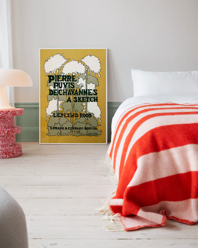 A cozy bedroom features a vintage Pierre Puvis de Chavannes Poster propped against the wall. A white bed is adorned with a red and white striped blanket, while the warm glow of a small lamp illuminates from a nearby table.
