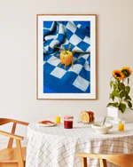 A dining area with a checked tablecloth and wooden chairs showcases juice, bread, and jam. The playful touch comes from the Little Visitor Poster, depicting a bell pepper as the sun on a checkered cloth. A sunflower plant completes this bright and inviting setting.