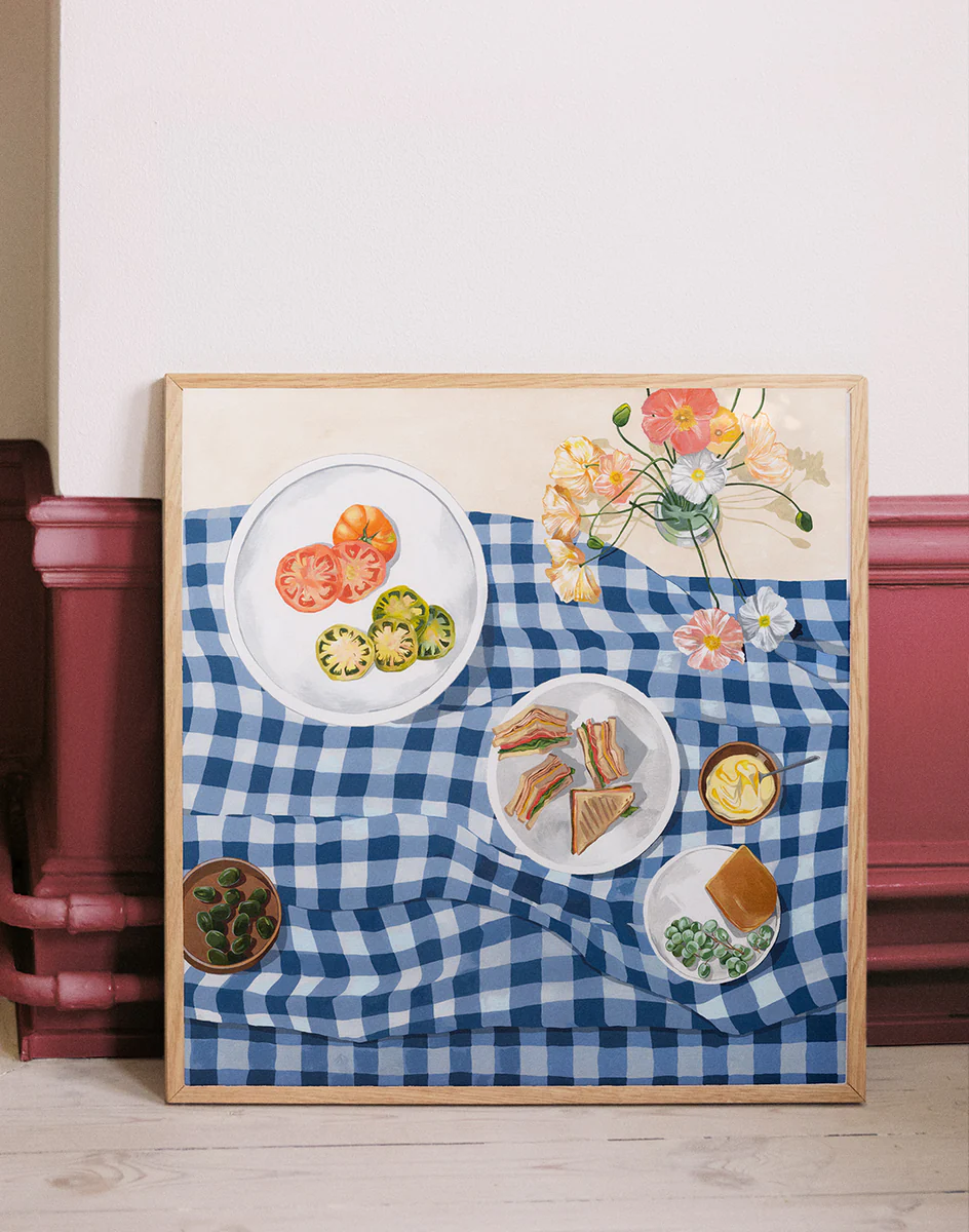 The Poppies and Picnics Poster features a painting of a blue and white checkered tablecloth with sliced tomatoes, sandwiches, and leafy greens. A vibrant floral arrangement completes the summer romance scene. Perfect for adding charm to your kitchen wall, it leans gracefully against a rustic backdrop.