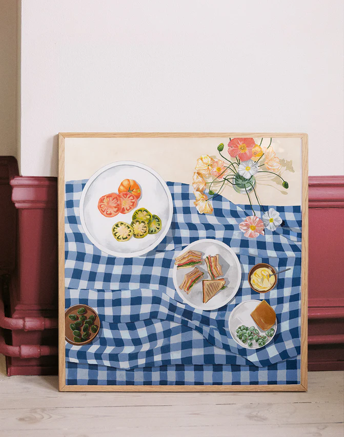 The Poppies and Picnics Poster features a painting of a blue and white checkered tablecloth with sliced tomatoes, sandwiches, and leafy greens. A vibrant floral arrangement completes the summer romance scene. Perfect for adding charm to your kitchen wall, it leans gracefully against a rustic backdrop.