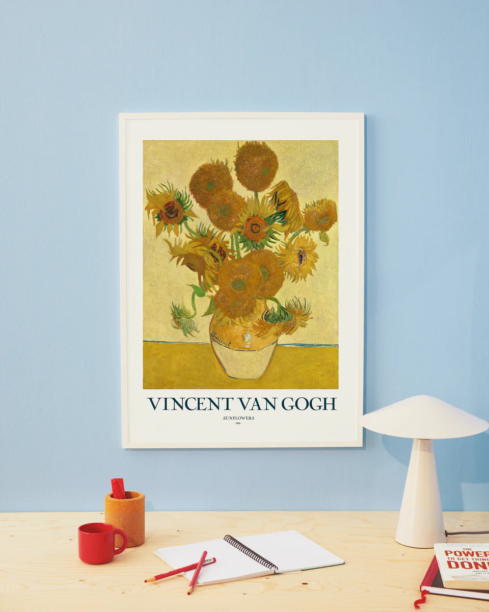 A framed Sunflowers by Vincent van Gogh poster decorates a light blue wall above a wooden desk, complemented by a white lamp, red mug, notebook, and scattered pens.