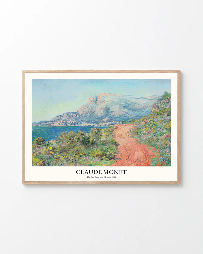 Framed print of Claude Monets The Corniche of Monaco showcasing Impressionist elegance, capturing a scenic landscape with a red dirt path, lush greenery, and coastal mountains against a white wall. This artwork beautifully reflects the charm of the French Riviera.