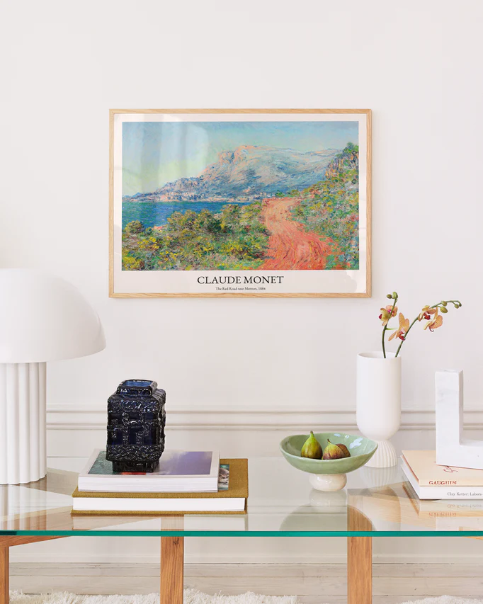 A modern living room features Impressionist wall decor with The Corniche of Monaco by Claude Monet Poster on a white wall. Below, a glass table holds stacked books, a green bowl of pears, a ceramic vase filled with flowers, and a black decorative object. A white lamp softly lights from the left.