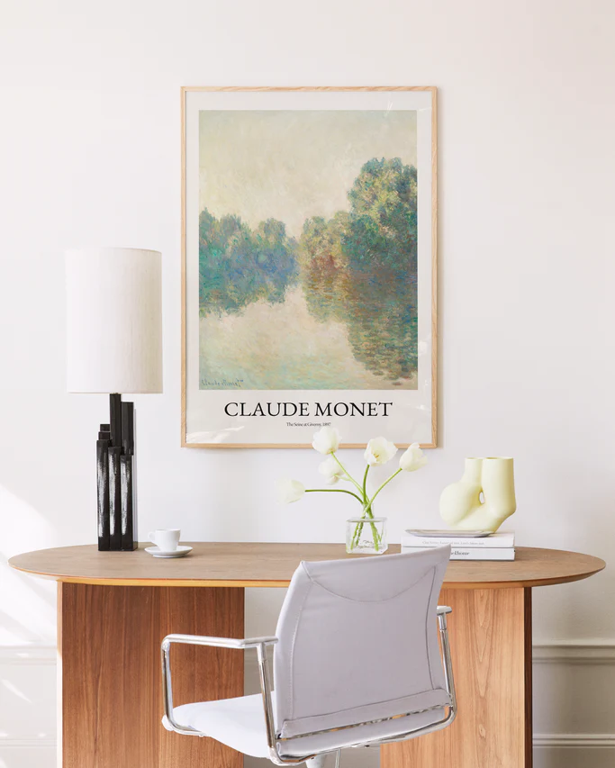 A minimalist office setup includes a wooden desk and a white chair. A lamp and a vase with white flowers sit on the desk, while The Seine at Giverny by Claude Monet poster evokes the serene beauty of the French countryside.