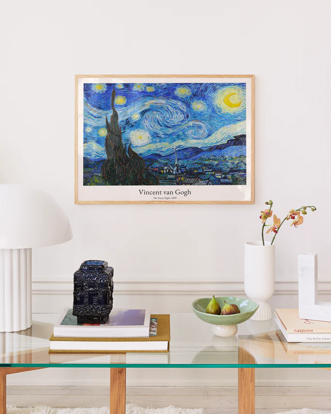 A stylish room features a Van Goghs The Starry Night poster on the wall. Below, a glass table displays a lamp, stacked books, a decorative box, a bowl with pears, and a small vase with flowers.