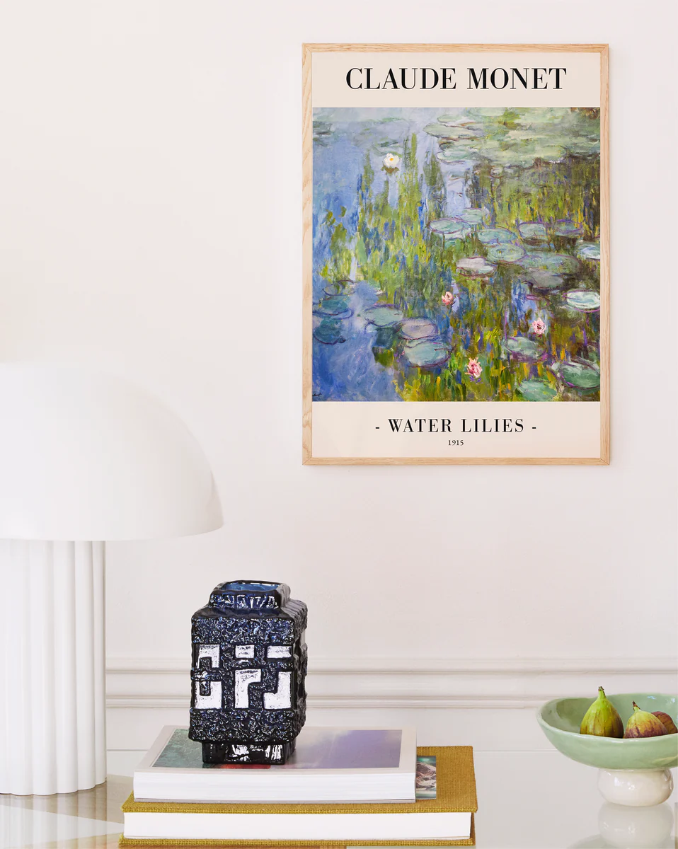 A soothing interior showcases a Claude Monet Water Lilies by Claude Monet 1915 Poster, elegantly framed in light wood. A white lamp and a small decorative box sit on stacked books, while figs in a bowl add nature to this French Impressionism-inspired scene.