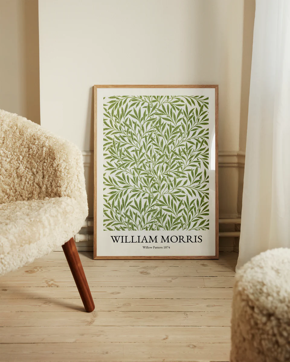 A framed William Morris Pattern 1874 Poster with a green leaf design leans against a wall. Beside it, a cream-colored textured chair is placed on a light wooden floor, bathed in soft natural light through the curtain, enhancing the scenes Victorian charm.