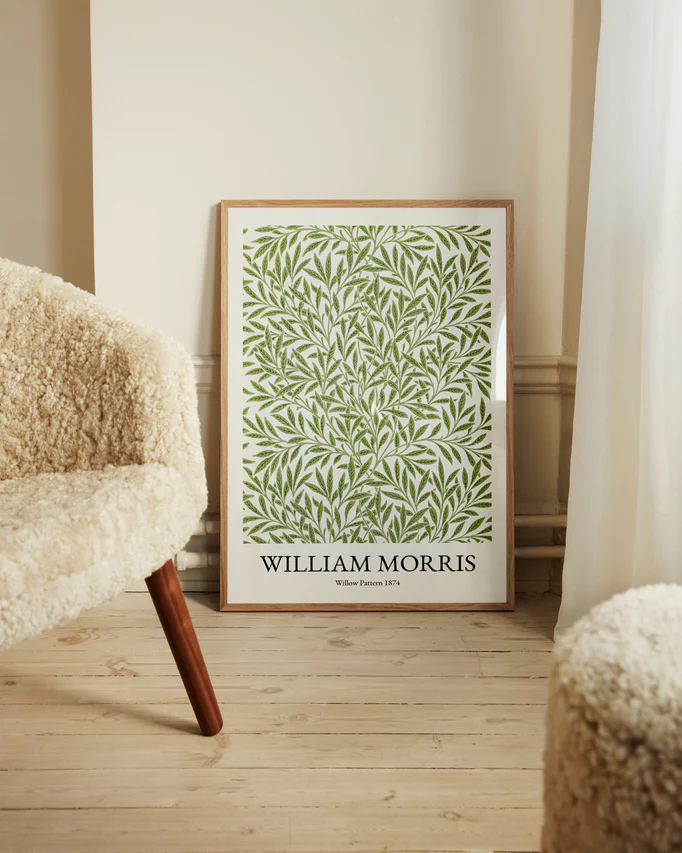 A framed William Morris Pattern 1874 Poster with a green leaf design leans against a wall. Beside it, a cream-colored textured chair is placed on a light wooden floor, bathed in soft natural light through the curtain, enhancing the scenes Victorian charm.
