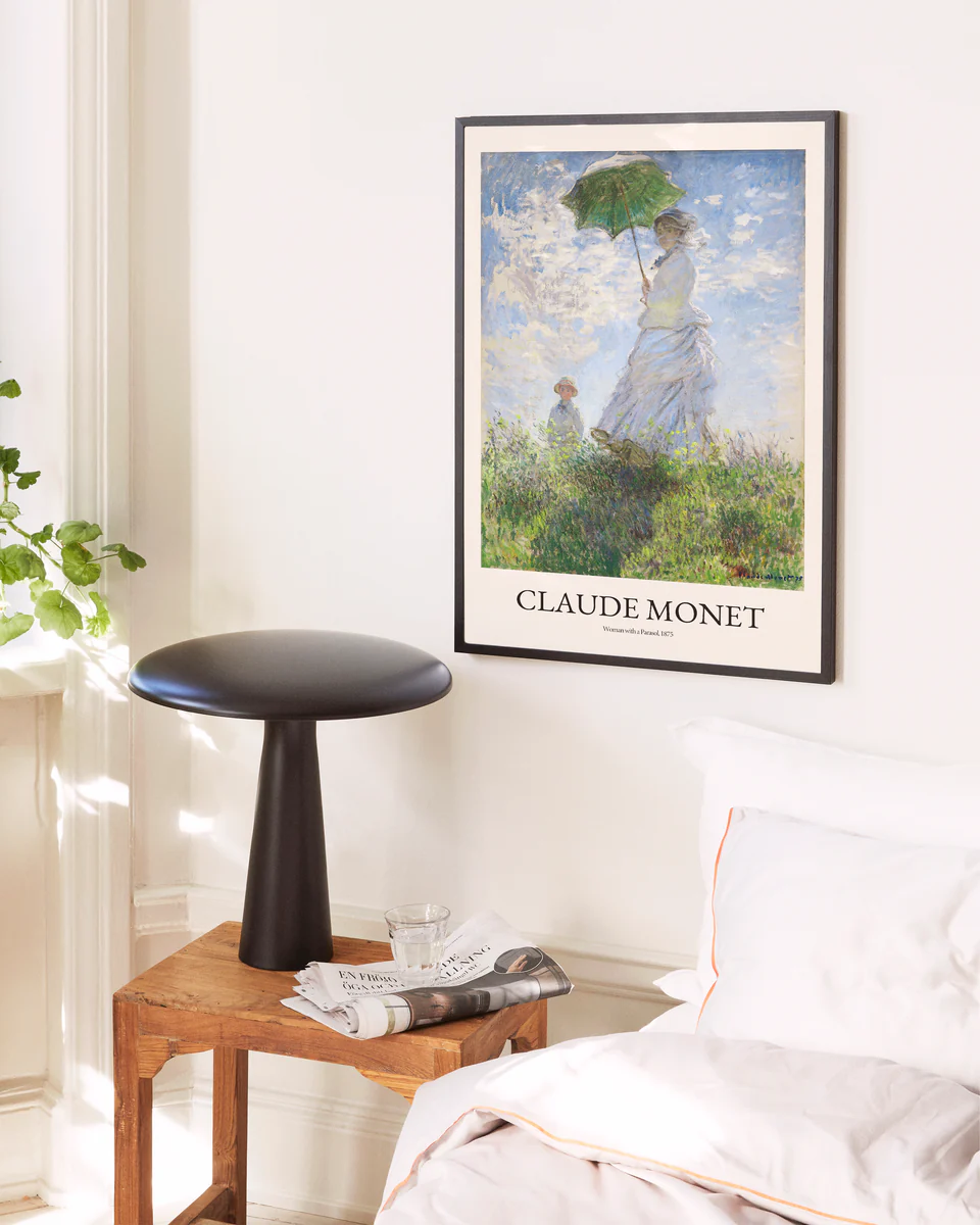 A cozy bedroom corner features a wooden nightstand holding a newspaper and glass. A modern black lamp is placed next to a bed with white linens. A Woman with a Parasol by Claude Monet poster adorns the wall, while a green plant thrives in the soft natural light nearby.