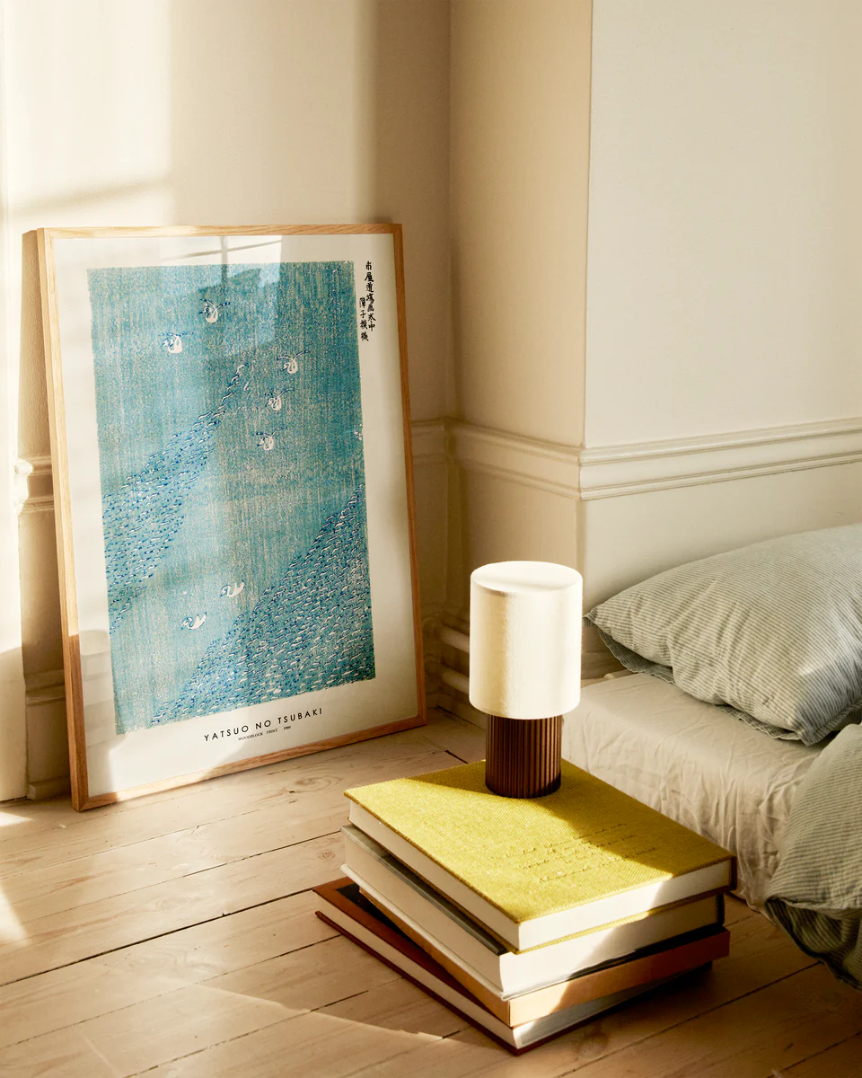 In a cozy bedroom corner, soft sunlight streams in. The Woodblock Print Blue by Taguchi Tomoki Poster leans against the wall beside a bed with light gray sheets. A small lamp rests on a stack of books, with one bright yellow, enhancing the home décor on the wooden floor.