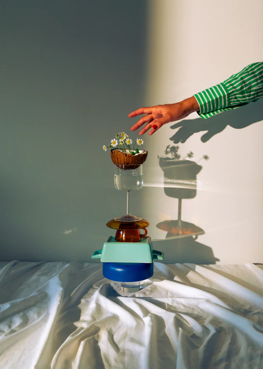 Small bowls, a wine glass delicately balanced atop each other, and at the top, a half coconut adorned with flowers. A hand reaching in for the blooms.