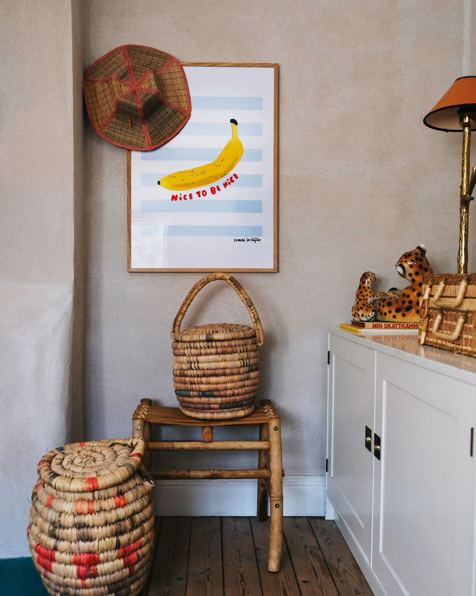 Poster Banana in an oak frame hanging in a children's room above a wooden stool. To the left of the oakframe hangs a cute little straw hat.
