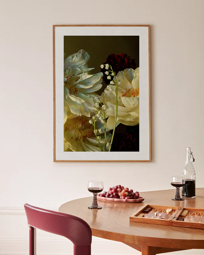 A room with a wooden table holding two glasses of red wine, a grape-filled plate, and a backgammon set. The Peony & Lily of the Valley Poster hangs above. A bottle of water is also on the table.