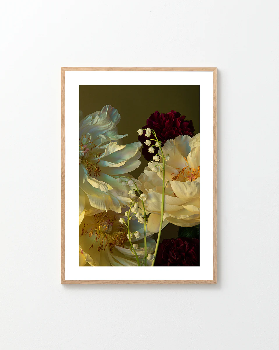 A framed Peony & Lily of the Valley Poster features a still life of white peonies, red carnations, and delicate Lily of the Valley blossoms against a dark background, elegantly displayed on a light-colored wall.