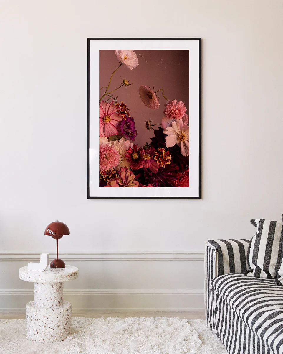 The Dreamscape Poster showcasing zinnias is displayed above a beige wall, enhancing the ambiance. A striped sofa sits beneath, paired with a round terrazzo side table holding a red lamp and book, crafting a cozy, modern living space.
