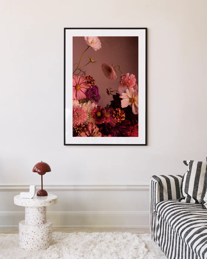 The Dreamscape Poster showcasing zinnias is displayed above a beige wall, enhancing the ambiance. A striped sofa sits beneath, paired with a round terrazzo side table holding a red lamp and book, crafting a cozy, modern living space.